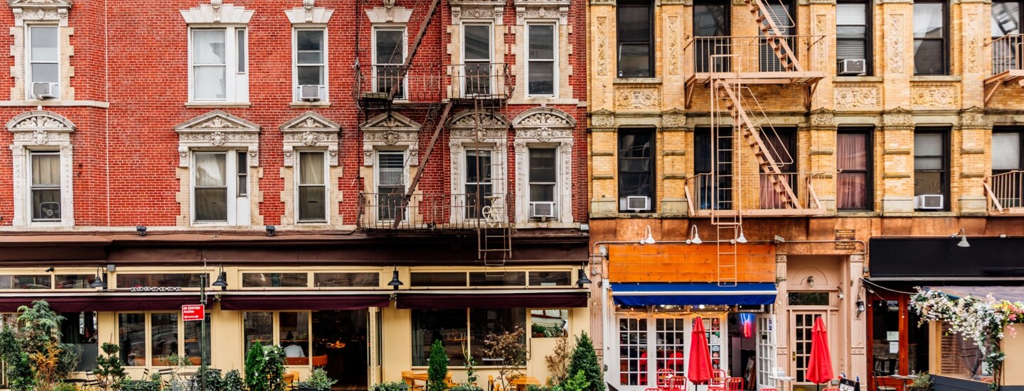 Geschlossene New Yorker Häuserfront mit Feuerleitern und Restaurants mit Außengastronomie.