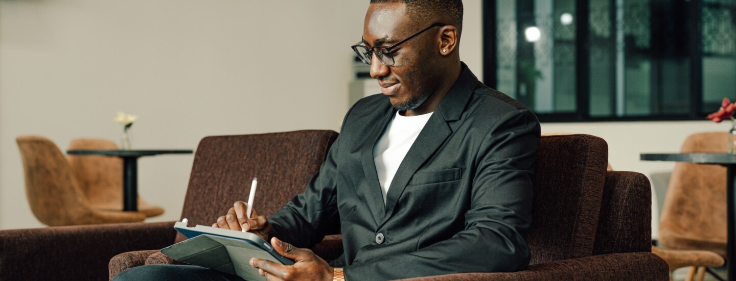 Ein Geschäftsmann arbeitet an einem Tablet auf einer Couch in einer Lounge, neben ihm steht ein Koffer.