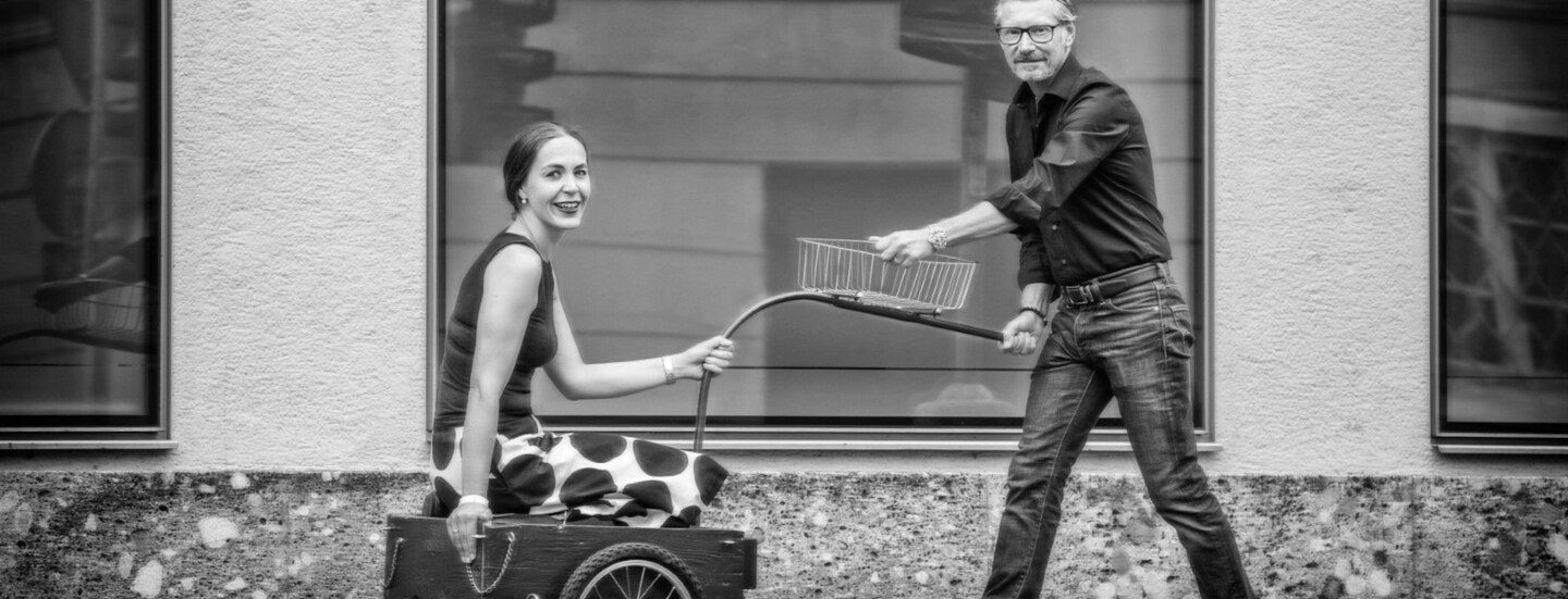 Ein Mann zieht eine lächelnde Frau in einem Fahrradanhänger auf einem Gehweg vor einer Fensterfront.