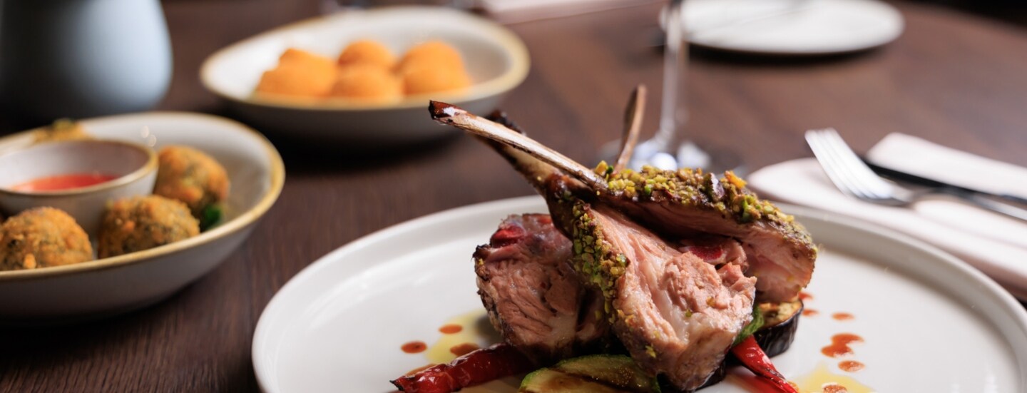 Gegrilltes Lammrippenkotelett, angerichtet auf einem weißen Teller, daneben Beilagen auf einem Tisch in einem Restaurant.