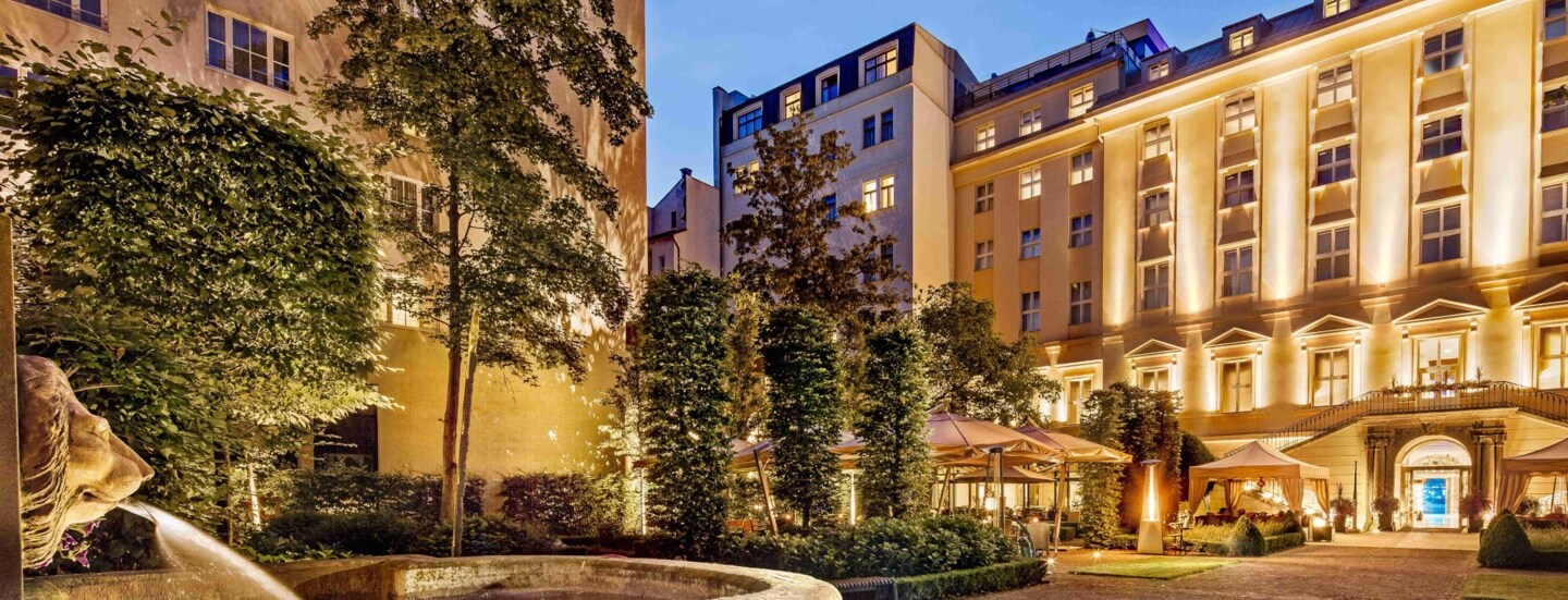 Imposantes, beleuchtetes Hotelgebäude mit barocker Fassade, im Vordergrund ein Springbrunnen mit Löwenkopf.
