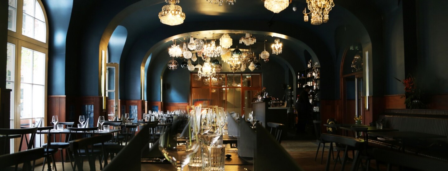 Gastraum des Restaurant AUGUSTIN in Köln mit eingedecktem Tisch im Mittelpunkt und Fokus auf der Deckenbeleuchtung aus vielen verschiedenen Kronleuchtern.