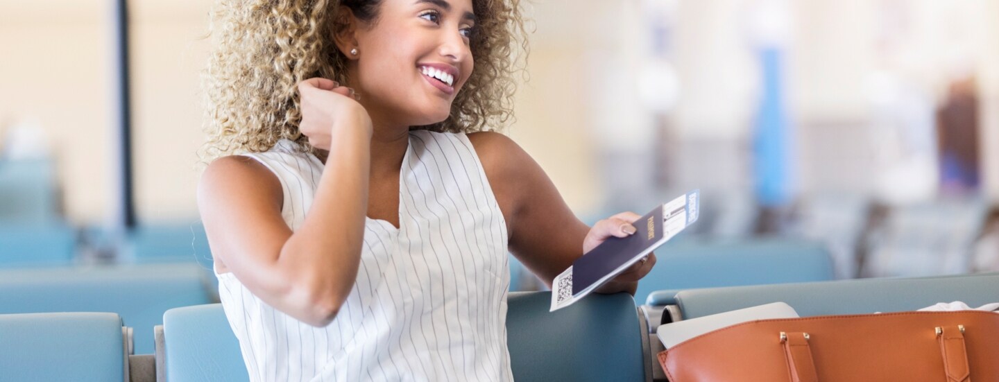 Eine junge, lächelnde Frau sitz mit Reisepass in der Hand in einer Flughafenlounge.