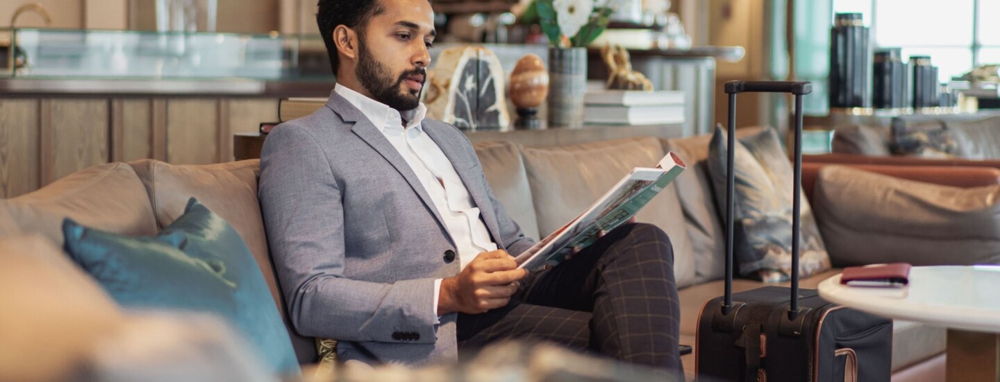 Ein elegant gekleideter Mann liest eine Zeitschrift auf einem Sofa in einer Flughafenlounge.