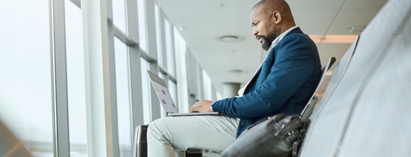 Ein Mann, der am Flughafen am Fenster sitzt und einen aufgeklappten Laptop auf dem Schoß hat.