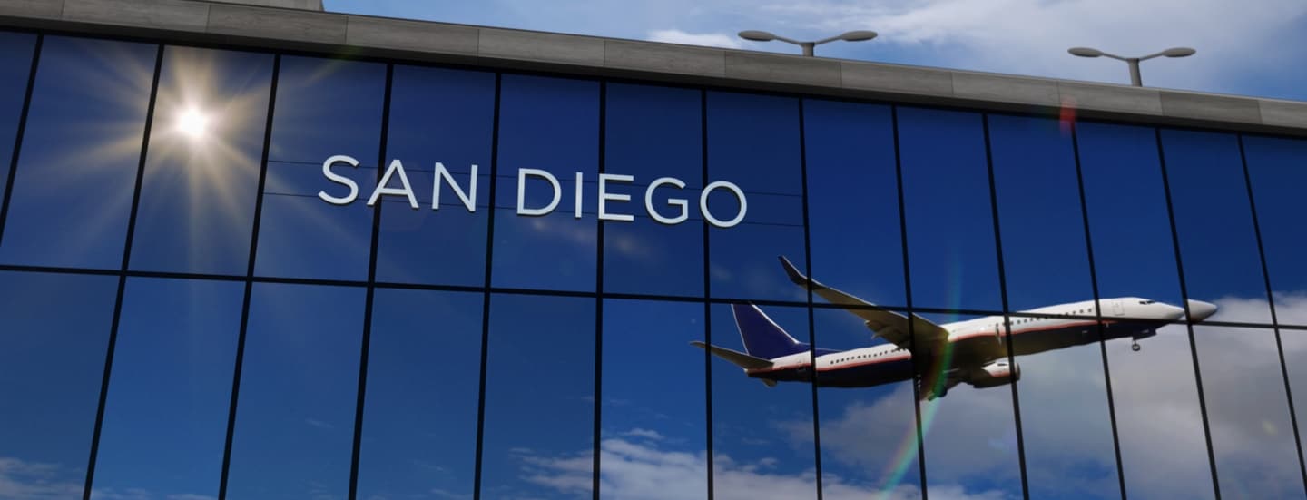 Flugzeug beim Abflug, das sich in den verspiegelten Fenstern einer Flughafenhalle spiegelt, auf denen die Aufschrift „San Diego“ zu lesen ist.