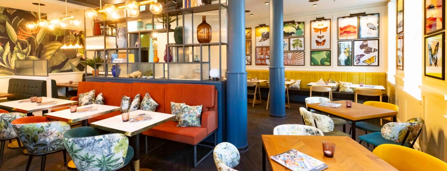 Innenraum des modern eingerichteten ALEX Restaurants an der Frauenkirche in Dresden, das mit großen Leuchten und einer Wandtapete mit Pflanzenmuster dekoriert ist.