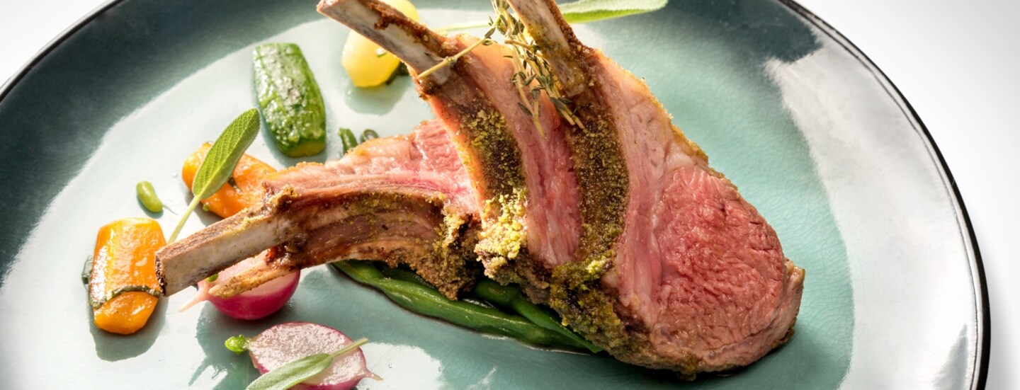 Lammkarree mit Kräuterkruste an Gemüse auf einem türkisfarbenen Teller.