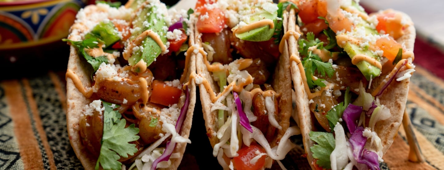 Drei aufgereihte Tacos, die mit Shrimps, Avocado, Koriander und mehr gefüllt und mit Soße angerichtet sind.