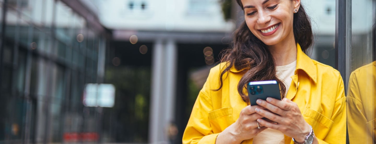 Eine Frau im gelben Hemd lehnt an einer Außenfassade und schaut lächelnd auf das Smartphone in ihren Händen.