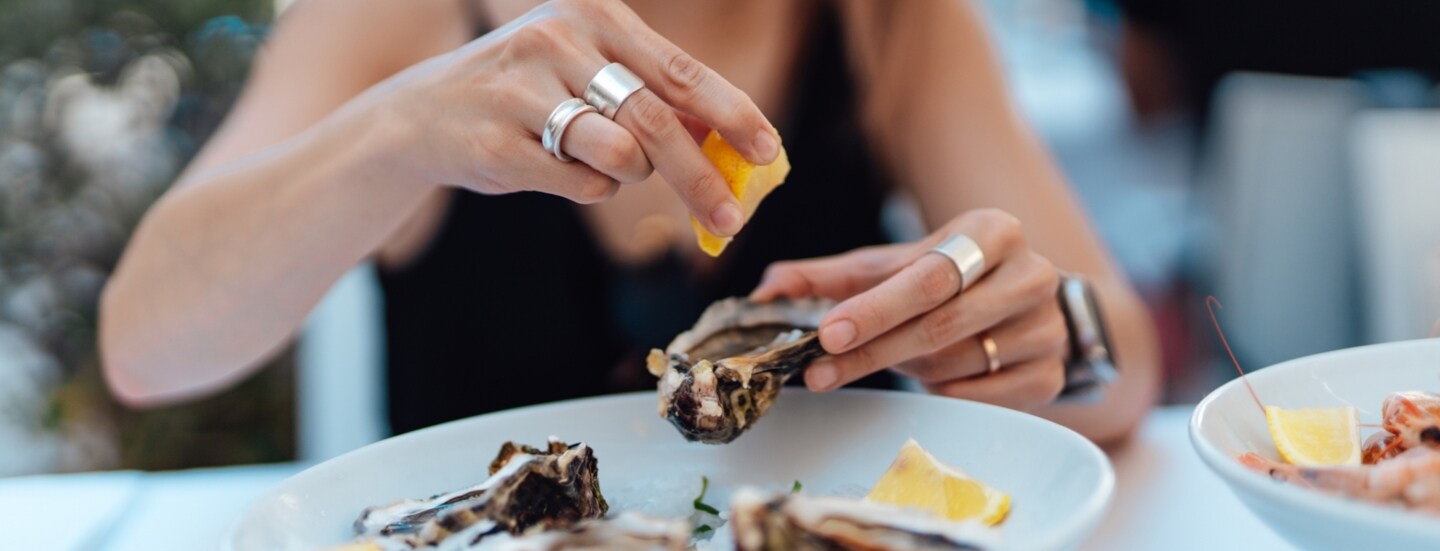 Eine unkenntliche Frau sitzt an einem Tisch, auf dem ein weißer Teller mit Austern auf Eis und Zitronenscheiben steh. Sie hält eine Auster und presst eine Zitrone darüber aus.
