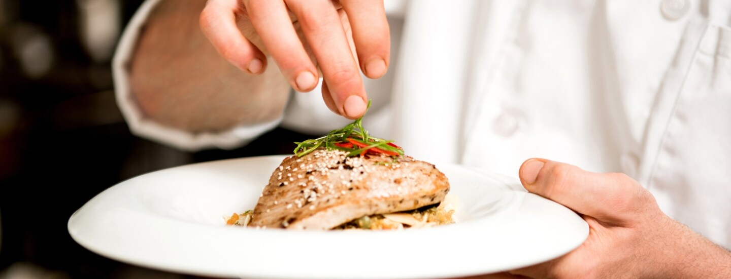 Nahaufnahme von einem weißen Teller mit einem Fleischgericht in den Händen einer Person in weißer Kochjacke.