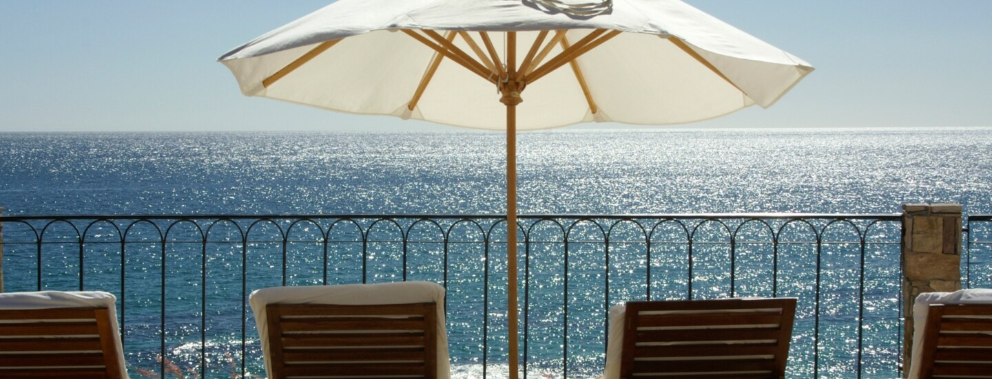 Terrasse und Geländer mit Blick aufs Meer, auf der mehrere Liegestühle und ein weißer Sonnenschirm stehen.