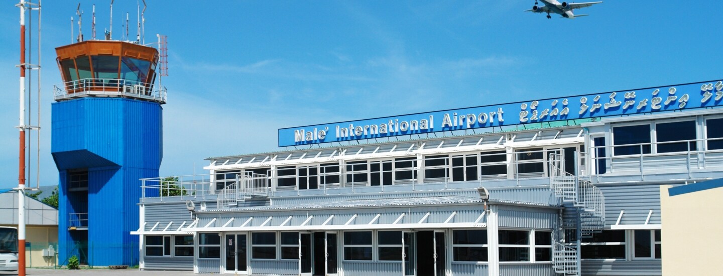 Ein Flugzeug fliegt am blauen Himmel über einen kleines Flughafengebäude mit blauem Kontrollturm.