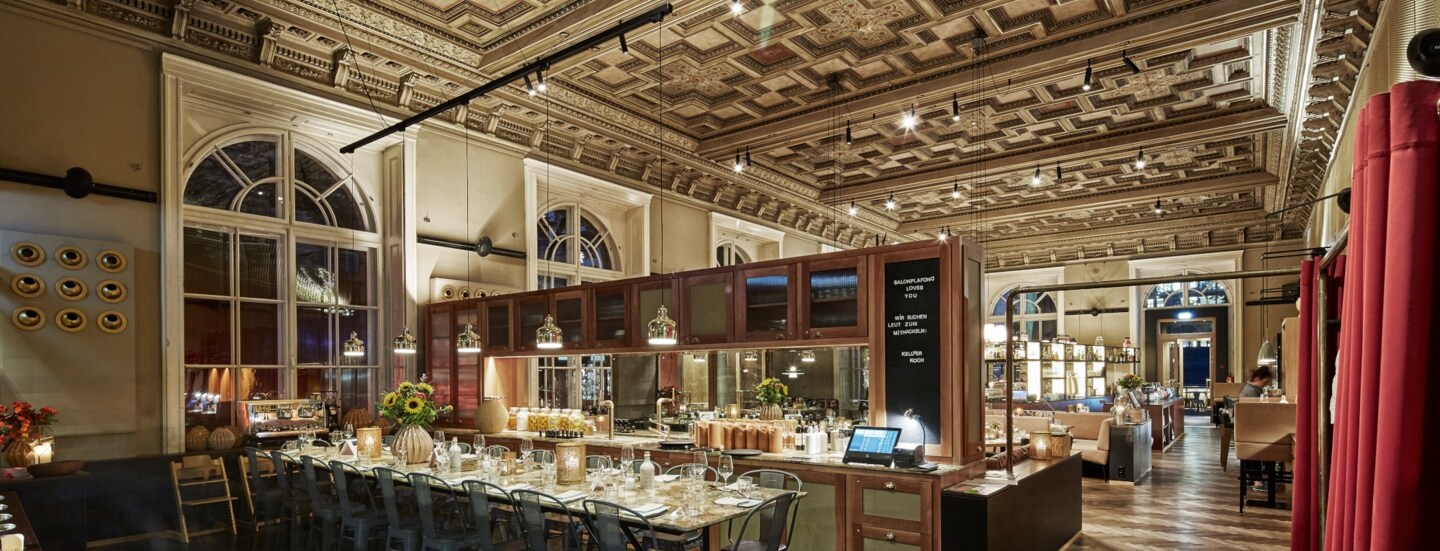 Restaurant mit einem langen, gedeckten Tisch, hohen Fenstern und Kassettendecke.