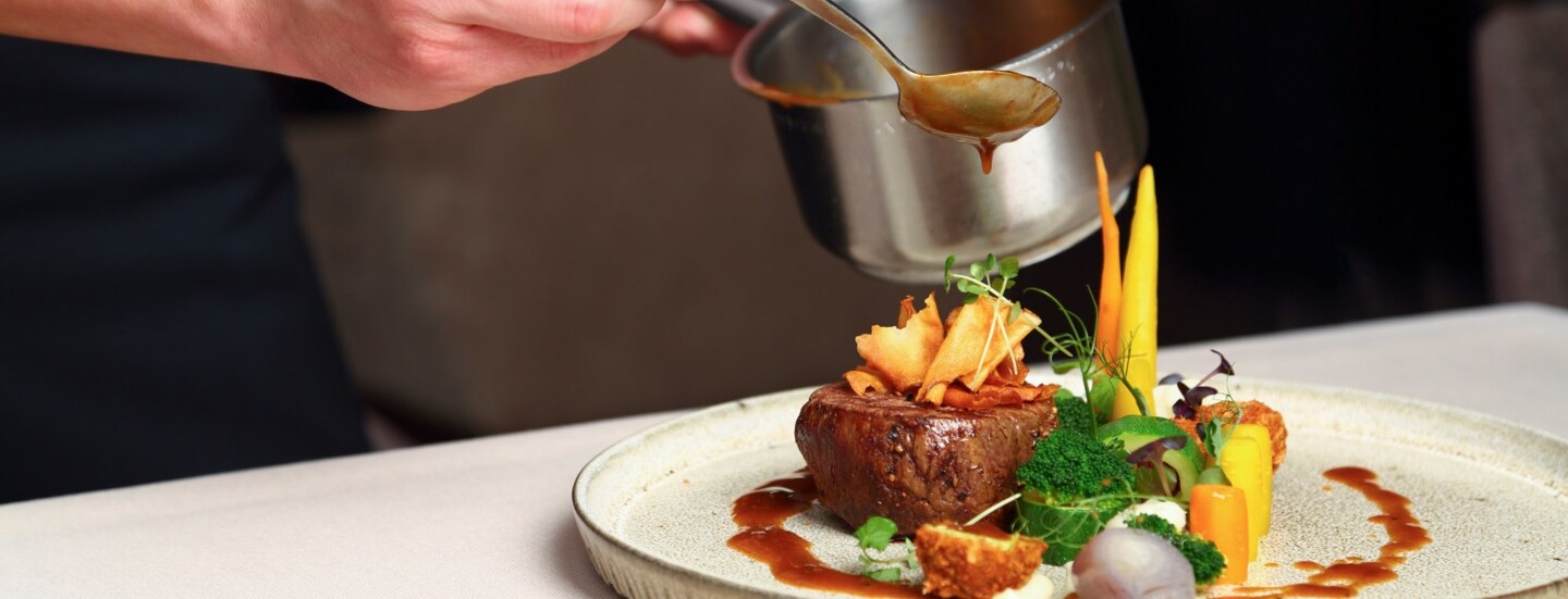 Eine Hand träufelt mit einem Löffel braune Soße auf ein Filetstück an Gemüse auf einem Teller.