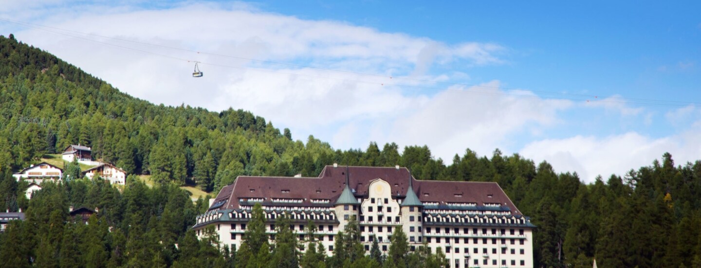 Ein Grandhotel mit zwei Türmen in einem Waldgebiet auf einem Berg, darüber fährt eine Seilbahn.