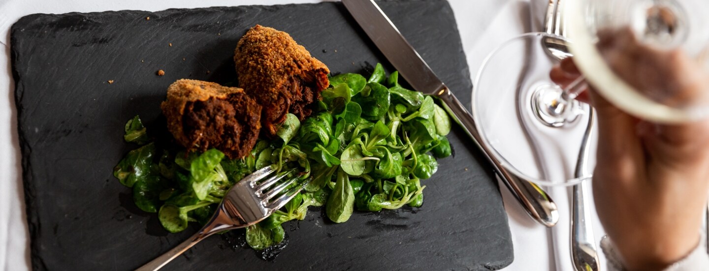 Aufsicht eines panierten Fleischgerichtes an Feldsalat auf einer Schieferplatte auf einem edel gedeckten Tisch, im Vordergrund eine Hand mit einem Glas Weißwein.