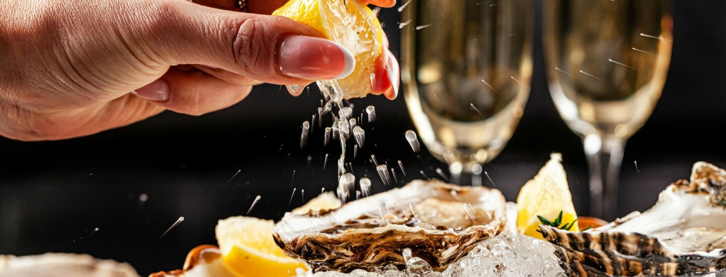 Eine Hand presst eine Zitrone über einem Teller mit Austern aus, dahinter zwei gefüllte Sektgläser.