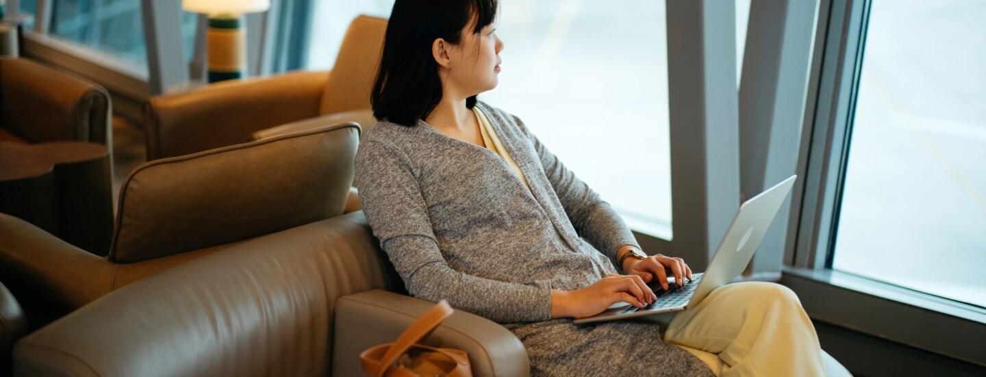Eine Frau sitzt mit Laptop in einem Ledersessel am Fenster einer Flughafen-Lounge und schaut aufs Rollfeld.