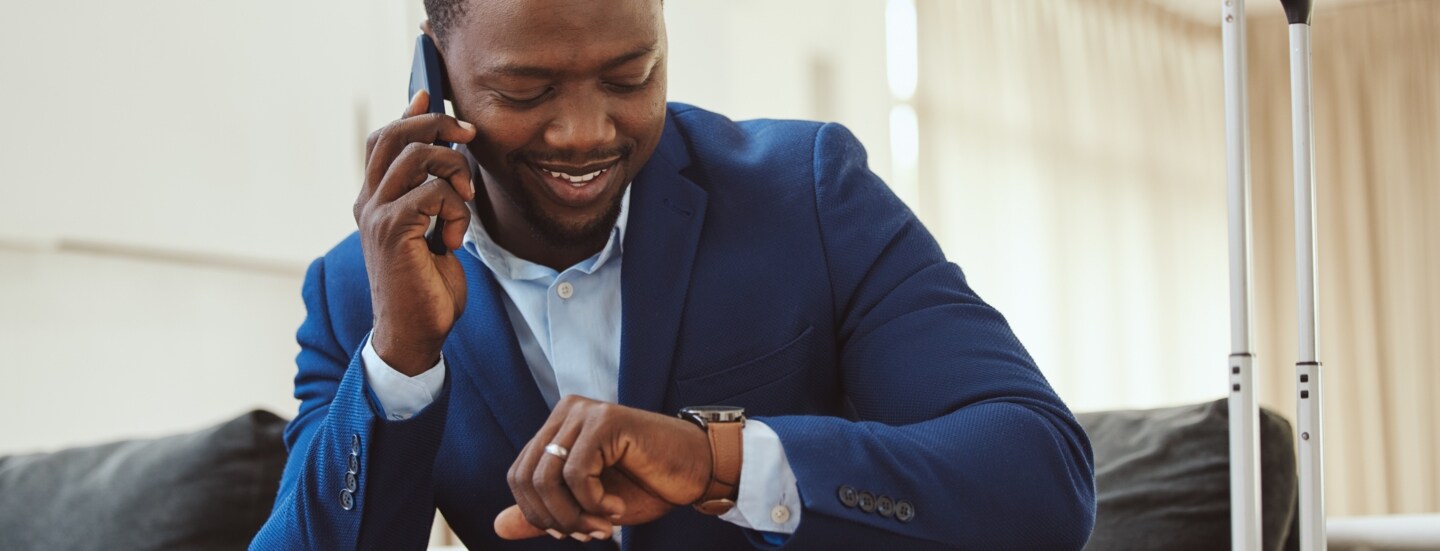 Ein lächelnder Geschäftsmann sitzt mit Smartphone am Ohr in einer Lounge und blickt auf seine Armbanduhr.