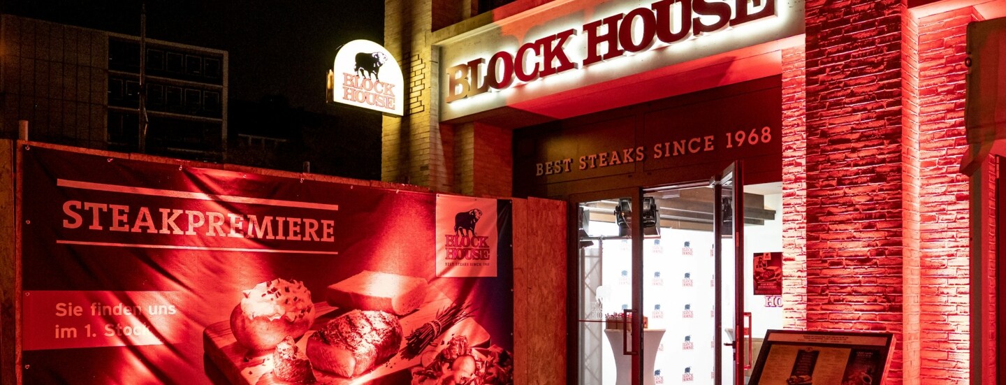 Rot beleuchteter Eingangsbereich eines BLOCK HOUSE Restaurants mit Werbebanner.