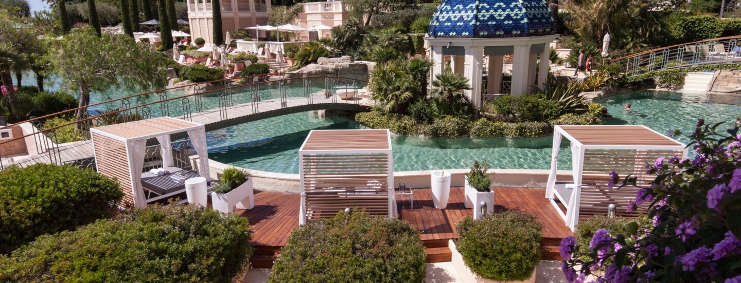 Ein lang gestreckter, mit Palmen, Blumen und Daybeds gesäumter Pool, über den zwei Brücken führen.