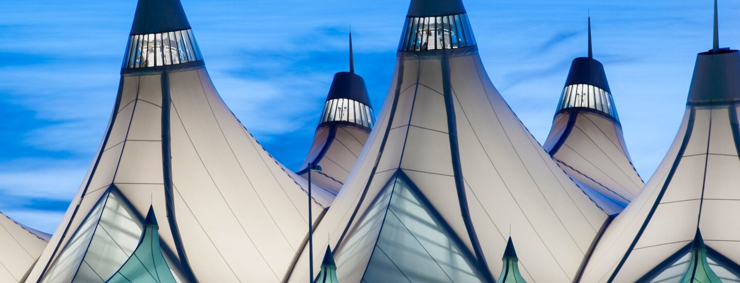 Beleuchtetes Gebäudedach aus Fibersegeltuch in konischen Formen vor blauem Himmel am Abend.