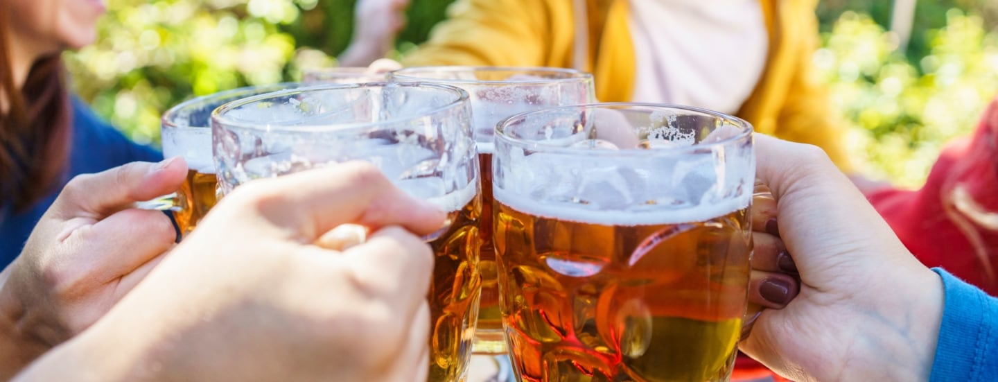 Nahaufnahme von vier gefüllten Bierkrügen aus Glas in den Händen von vier Personen beim Anstoßen an einem Tisch im Freien.