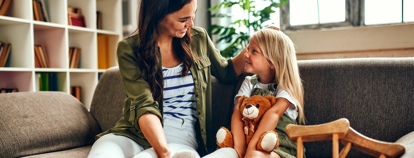 Ein kleines Mädchen mit eingegipstem Bein und Teddybär auf dem Schoß wird von ihrer Mutter auf dem Sofa getröstet.