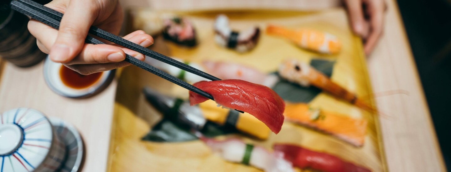 Eine Hand mit Stäbchen über einem Teller mit verschiedenen Sushi-Sorten.