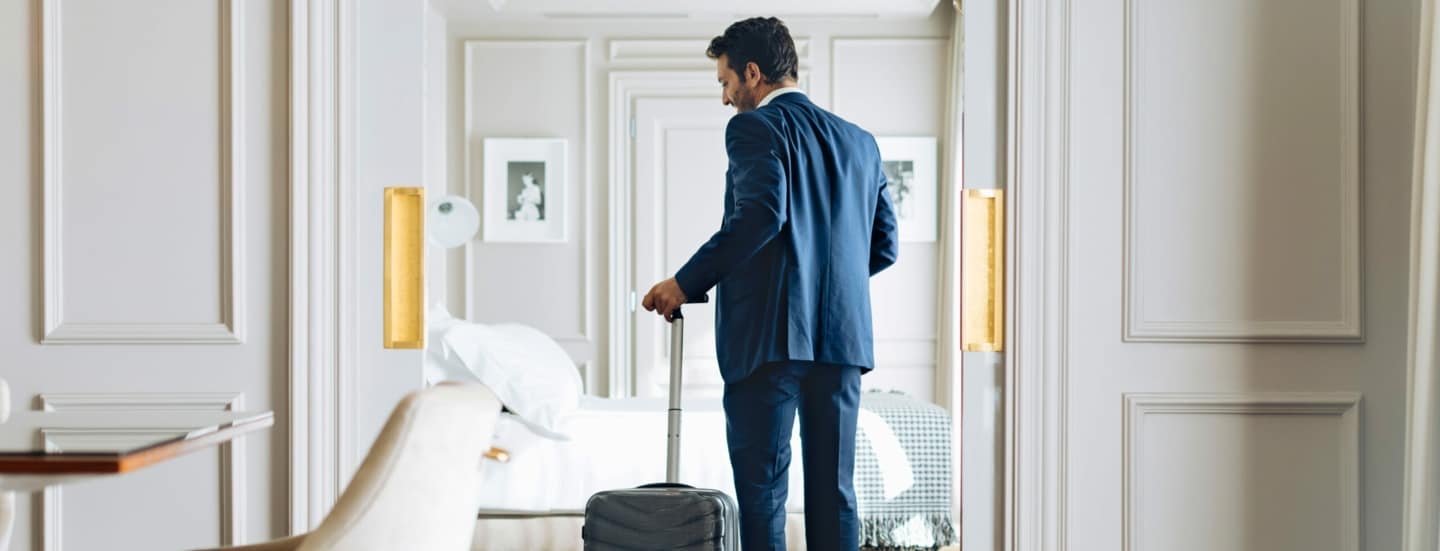 Mann im Anzug und einem Rollkoffer in der Hand betritt eine helle luxuriöse Hotel-Suite.