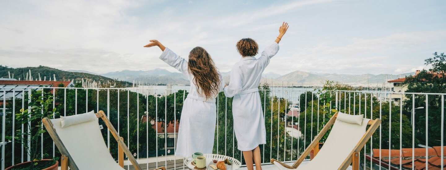 Zwei Menschen in weißen Bademänteln von hinten stehen auf einem Balkon, blicken ins Grüne und strecken freudevoll die Arme in die Luft.