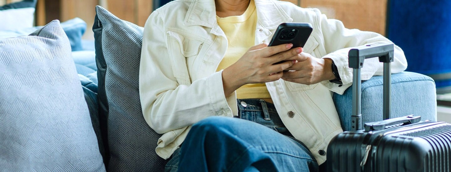 Eine junge asiatische Frau sitzt iauf einem Sessel in einer Lounge und schaut auf ihr Smartphone, neben ihr steht ein Hartschalenkoffer.