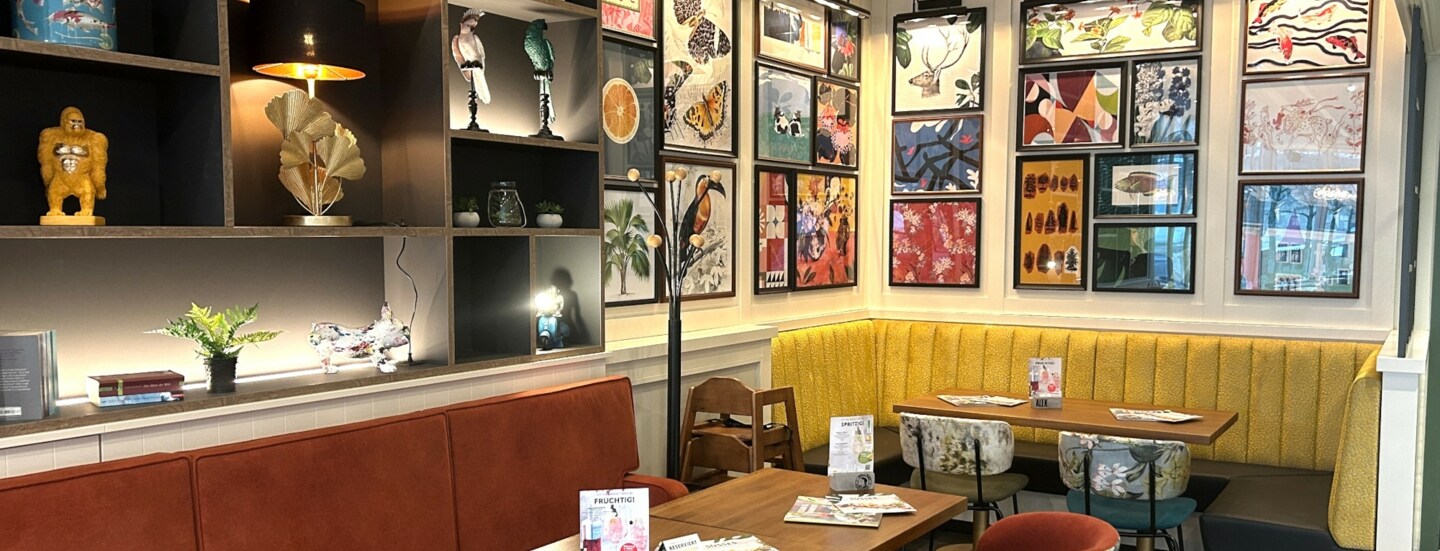 Gemütlicher Gastraum mit gelber Eckbank, bunten Stühlen und zahlreichen farbenfrohen Bildern an den Wänden.