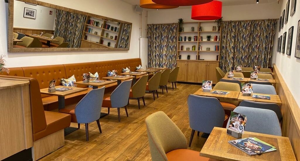 Gastraum mit Holzboden, bunten Stühlen, brauner Polsterbank, großen farbigen Deckenlampen und Bücherregal im Hintergrund.