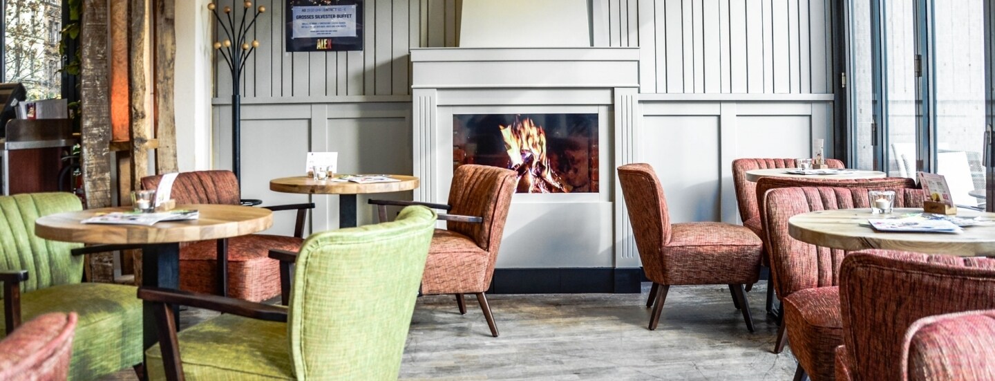 Gemütlicher Gastraum des ALEX in Wiesbaden mit loderndem Gaskamin, runden kleinen Tischen und bequemen Sesseln in Grün und Rot.