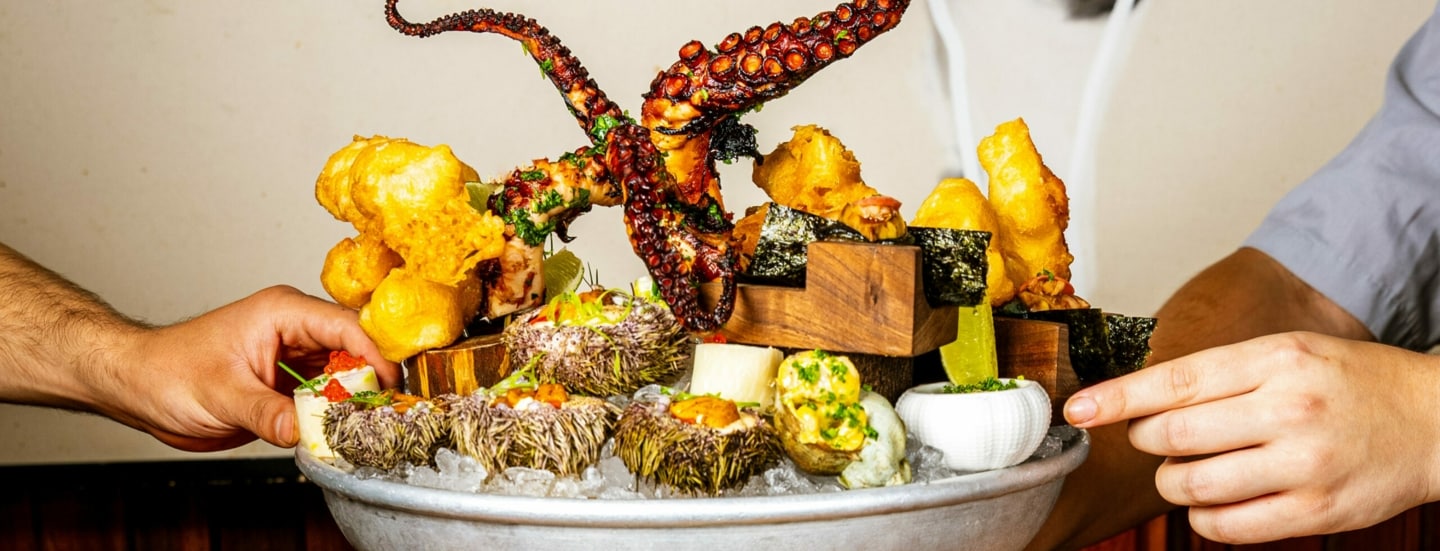 Hände drapieren auf einer tiefen Platte mit Eis diverse Speisen – von Tintenfisch über Frittiertes bis Kaviar.