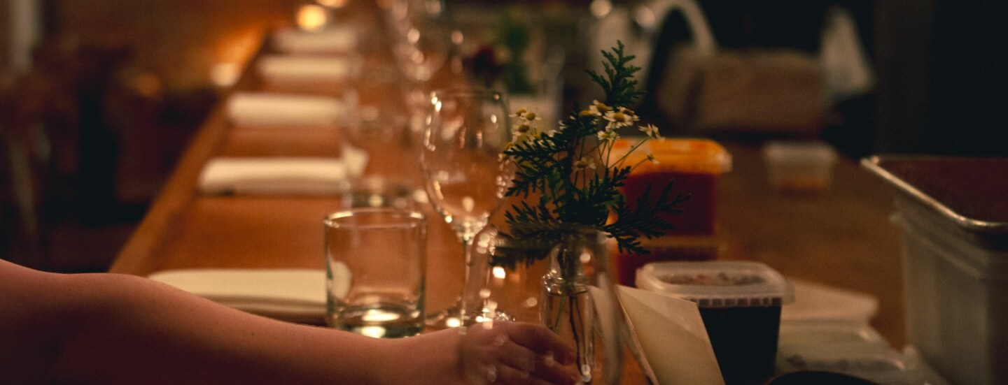 Gedeckter Holztresen bei schummrigen Licht, auf den eine Frauenhand eine kleine Blumenvase mit Kamille stellt.