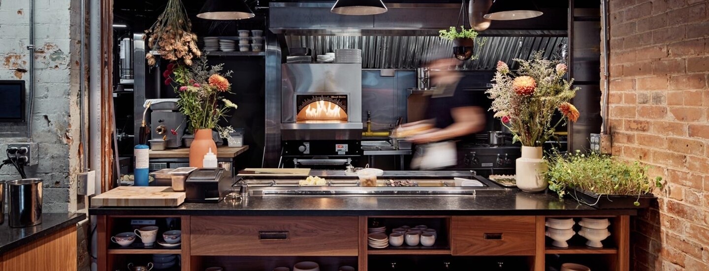 Blick in die Küche, wo im Ofen ein Feuer brennt und neben der unverputzten Backsteinwand eine Anrichte mit Blumensträußen und frischen Kräutern steht.