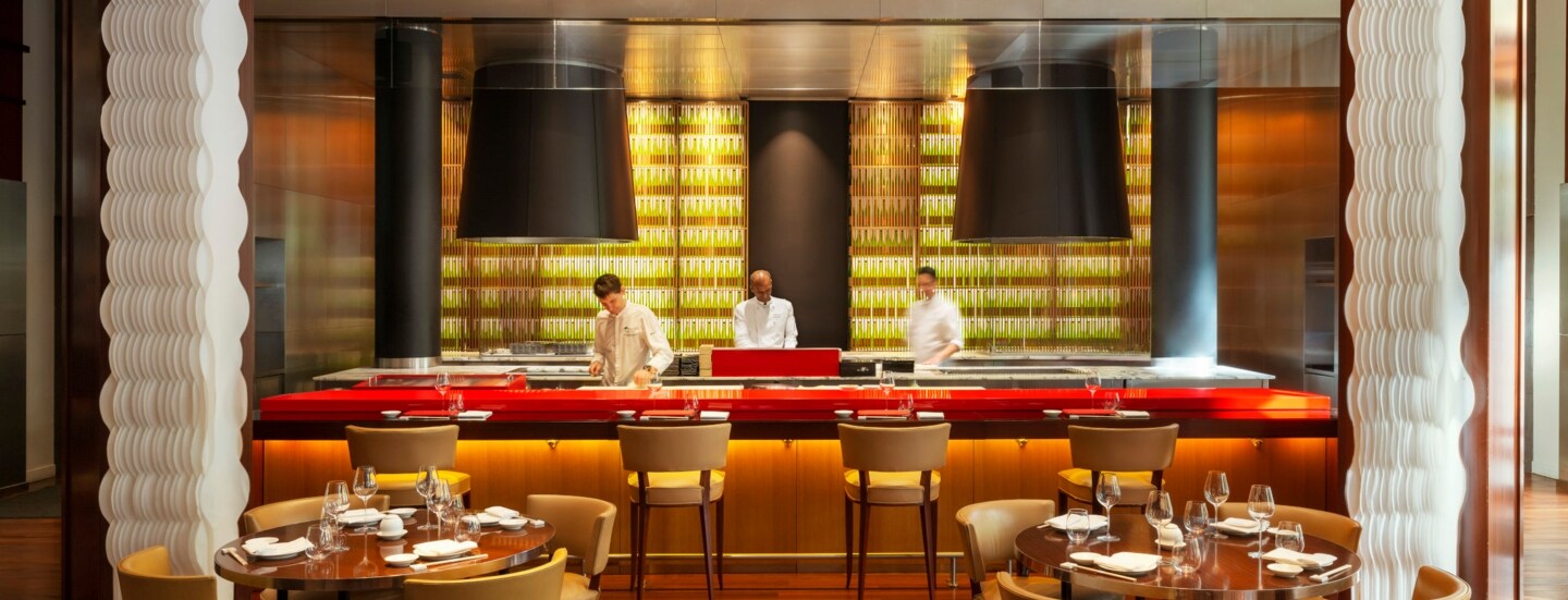 Interieur eines elegant-modernen Restaurants mir runden gedeckten Holztischen und offenem Tresen, in dem drei Köche stehen.