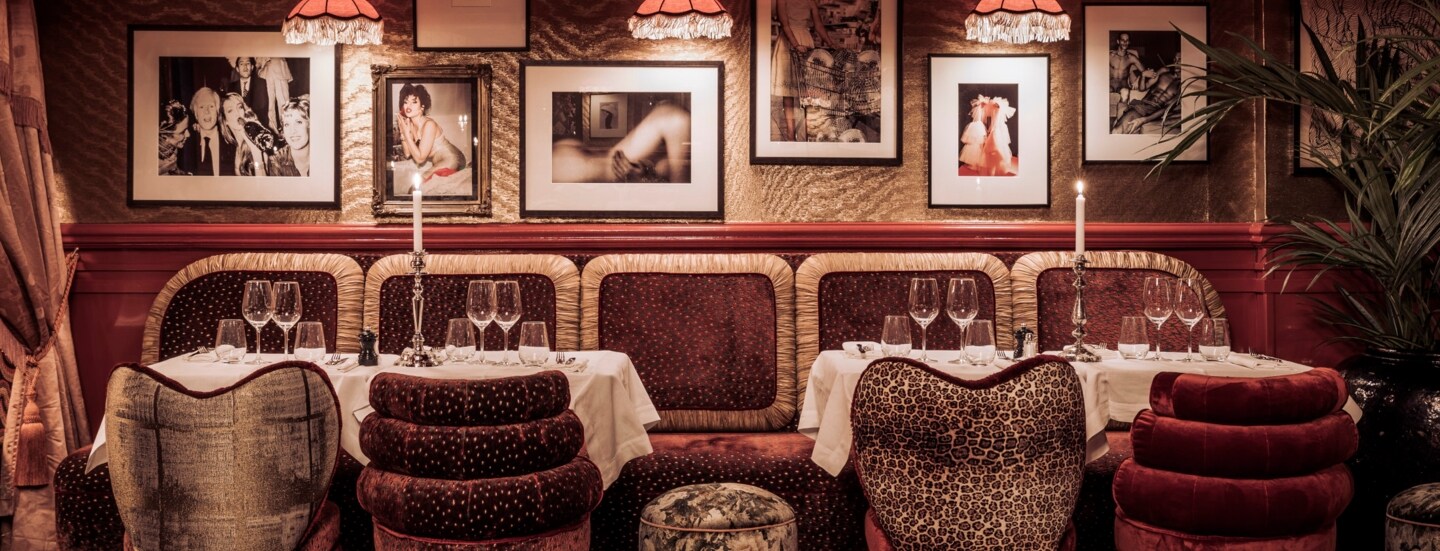 Gedeckte Tische in einem in dunklen Farben gehaltenem Interieur mit Sesseln und Teppich mit Animal-Muster sowie Fotografien an der Wand und Fransenlampen an der Decke.