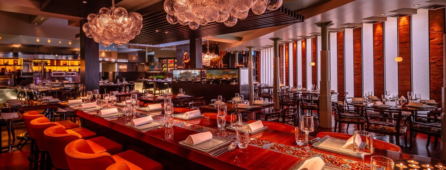 Opulent eingerichteter Speisesaal mit eingedeckter Holztafel und offener Küche.