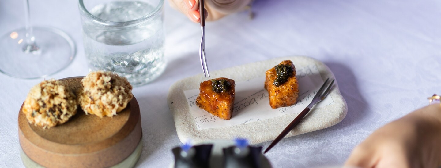 Eine Hand führt eine Gabel zu einem kunstvoll angerichteten Dessert auf einem weiß eingedeckten Restauranttisch.