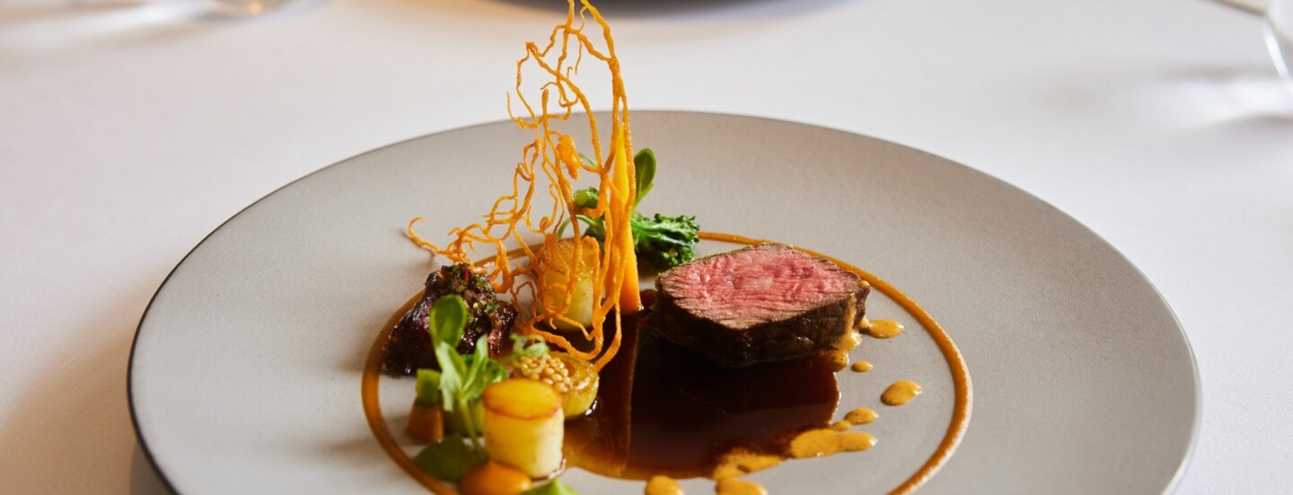 Kunstvoll arrangiertes Gericht mit einem Stück Fleisch auf einem großen weißen Teller auf einem weißen Tisch.