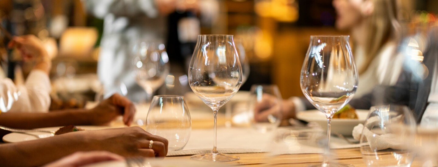 Weingläser auf einem gedeckten Holztisch in einem Restaurant mit Menschen im Hintergrund.