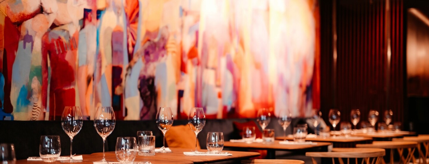 Mehrere eingedeckte Holztische vor einem bunten, großflächigem Wandbild.