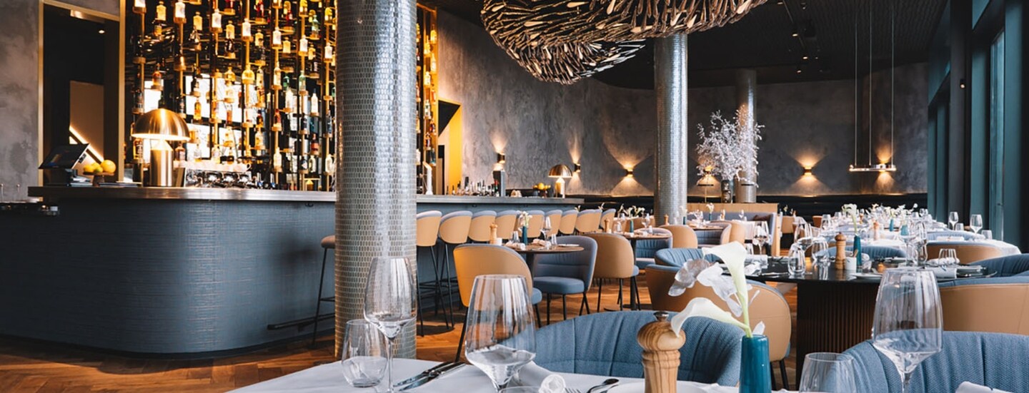 Stilvoller, modern eingerichteter Speisesaal mit Barbereich und eingedeckten Tischen unter einer abstrakten Deckenskulptur.