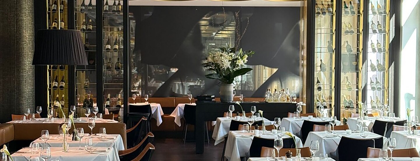 Eleganter Speiseraum mit weiß eingedeckten Tischen und goldfarbenen Deckenlampen vor einem schwarzen Wandbild.