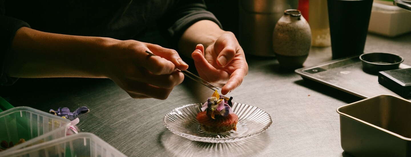 Zwei Hände garnieren eine edle Vorspeise mit Blüten auf einer Küchentheke.